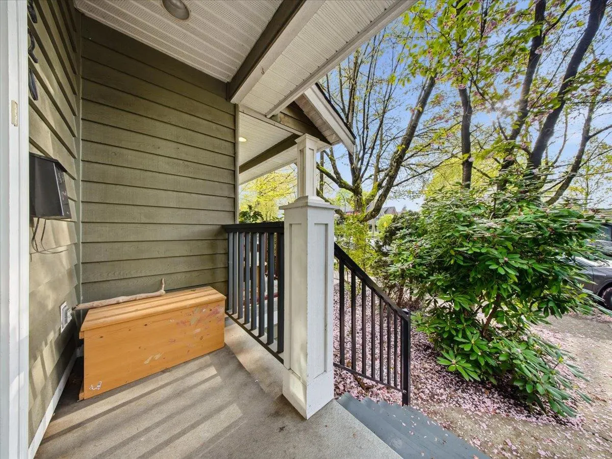 Covered porch with olive green siding, a wooden chest, and black railings leading to steps. Trees and foliage are visible.
