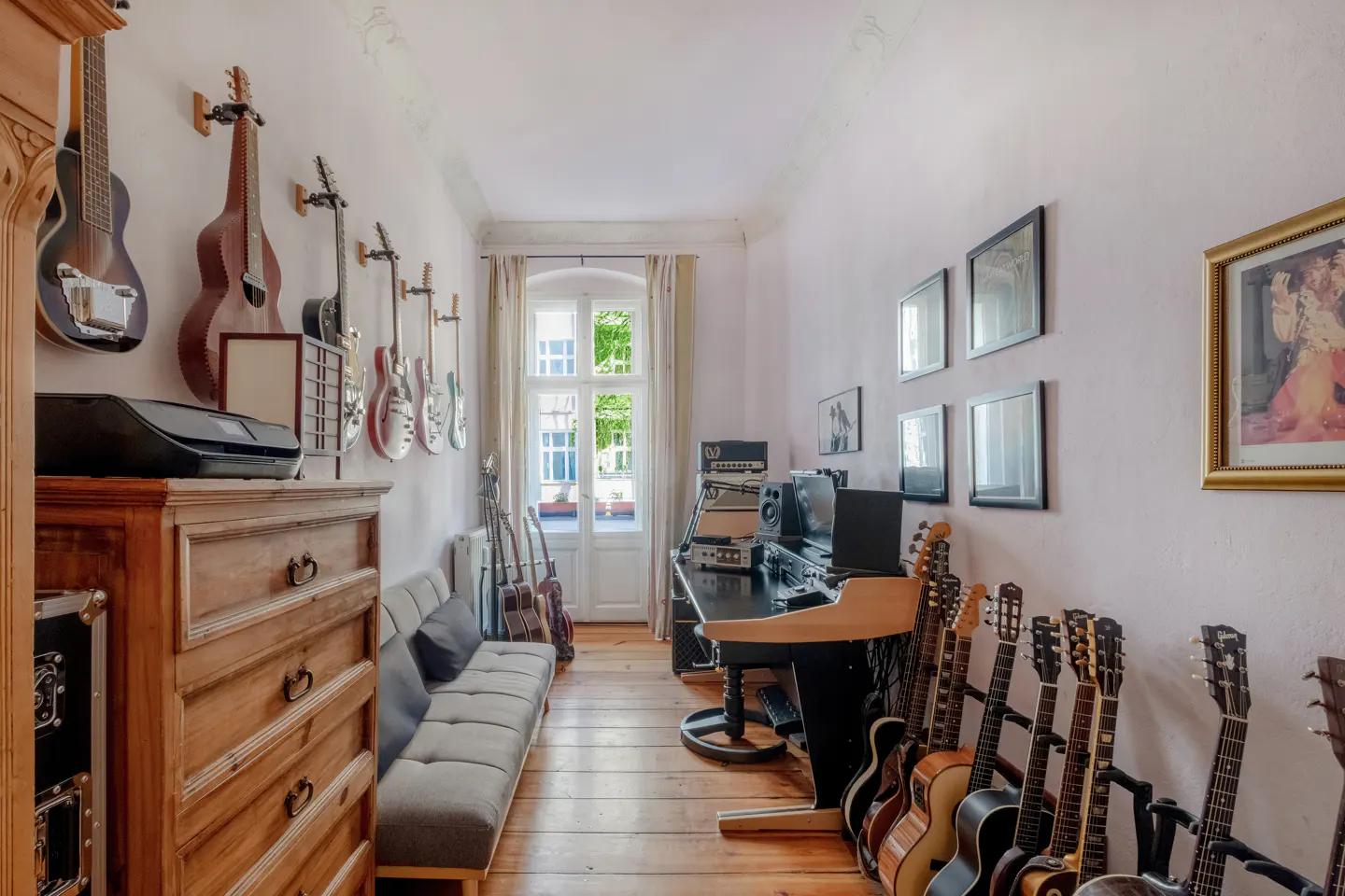 A music room with guitars on the walls, a sofa, a desk with recording equipment, and a window with natural light.