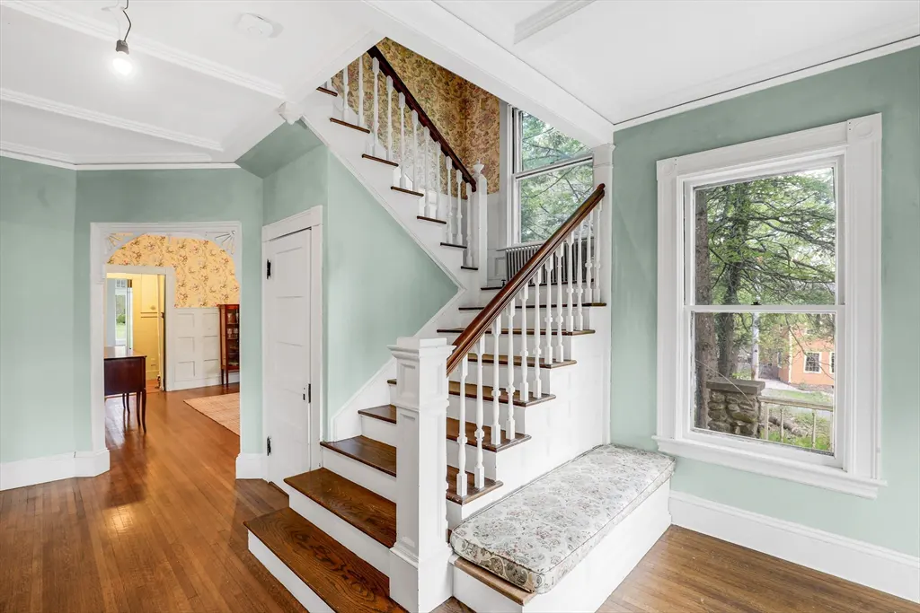 Interior view of a home's foyer with hardwood floors, a white staircase with a wooden banister, and a window seat. Walls are light green.