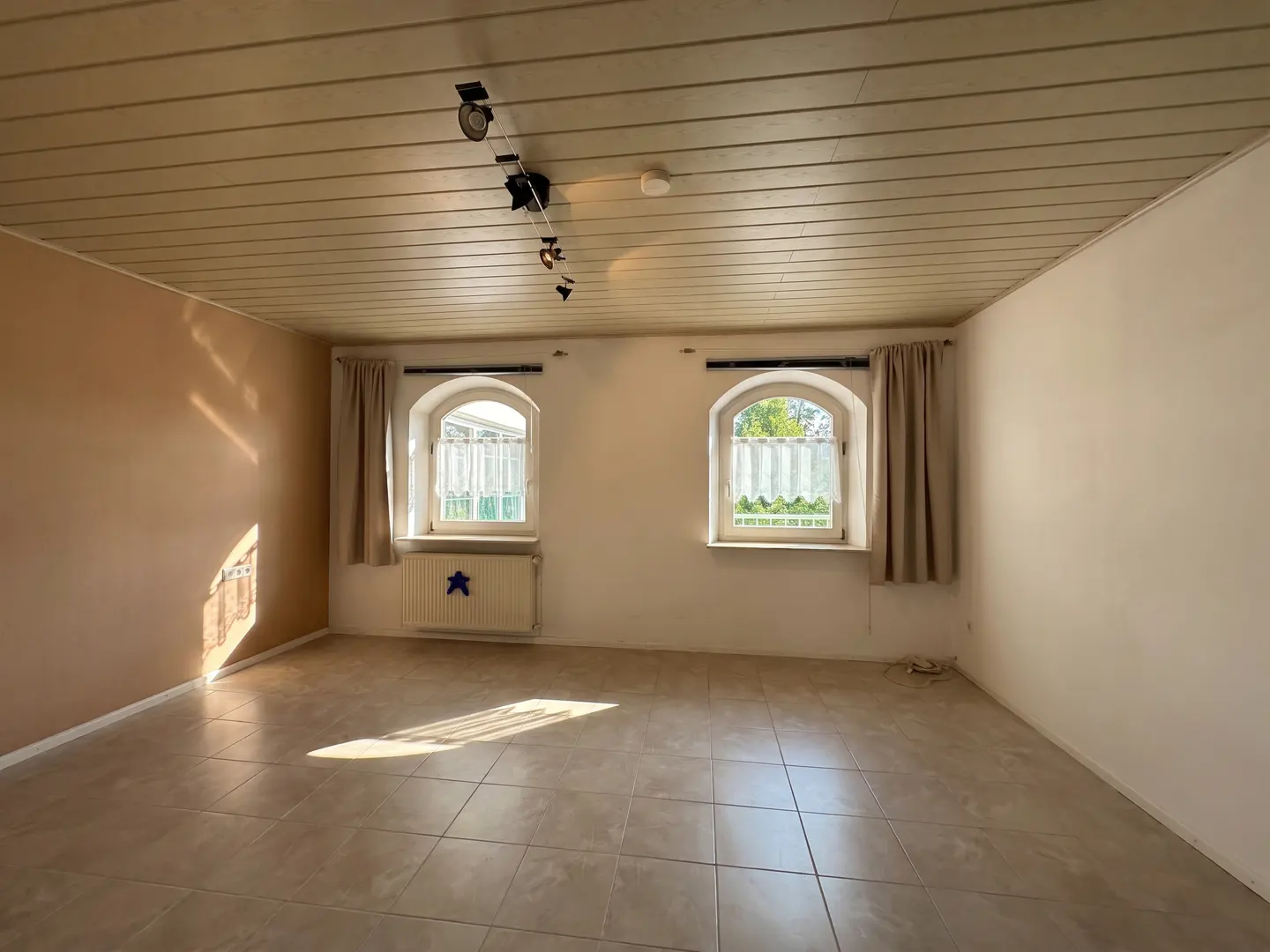 Empty room with beige tile floor, two arched windows with curtains, and a wood-paneled ceiling with track lighting.