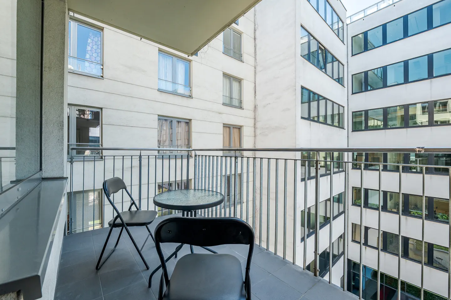 Balcony with two black chairs and a glass table, overlooking a white building with many windows.