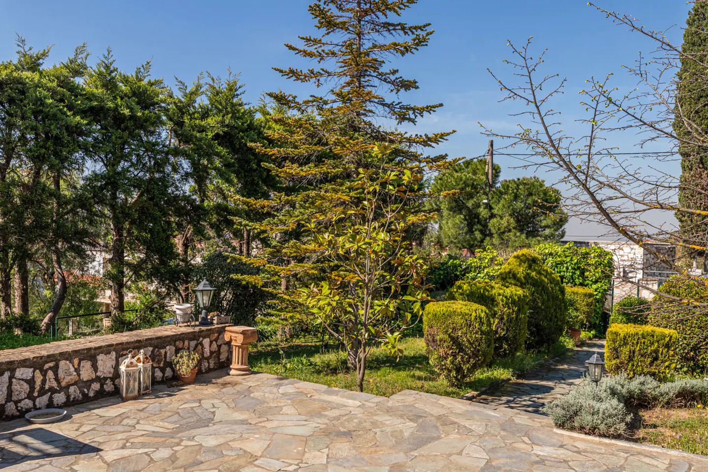 Stone patio with trees and shrubs. A stone wall with lanterns and a small column is visible. The sky is blue.