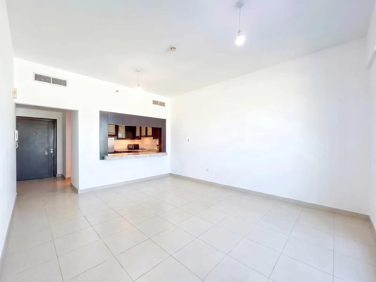 Empty living room with white walls, light tile floors, and a pass-through window looking into a dark wood kitchen. [AI-generated text]