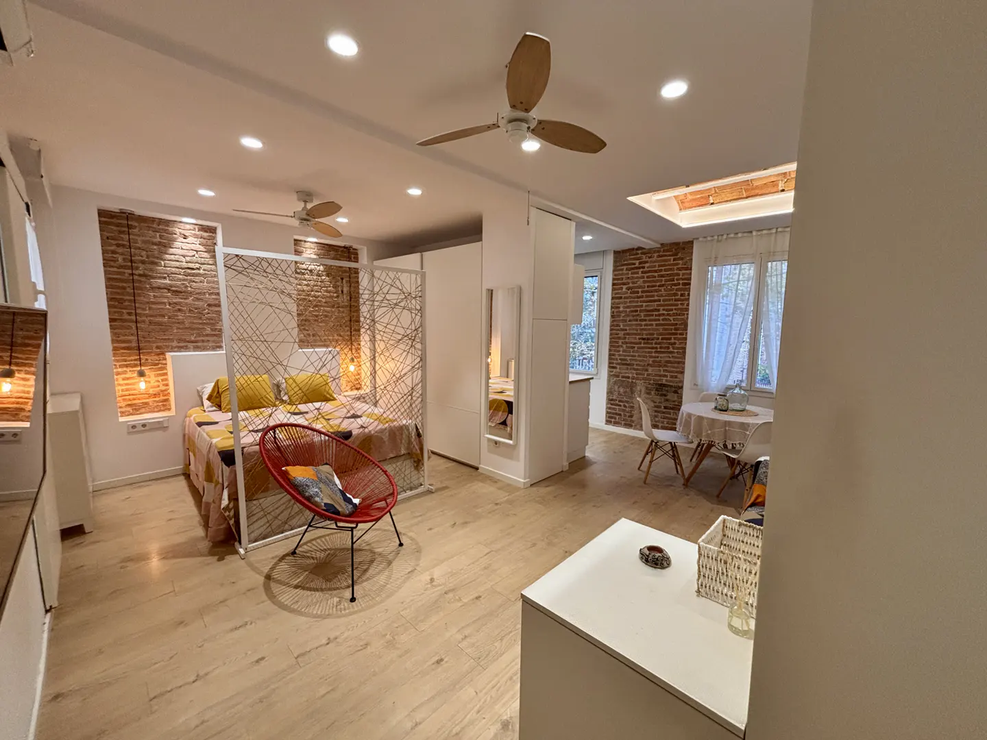 Modern studio apartment with a bed behind a decorative screen, a red accent chair, dining area, and exposed brick walls.
