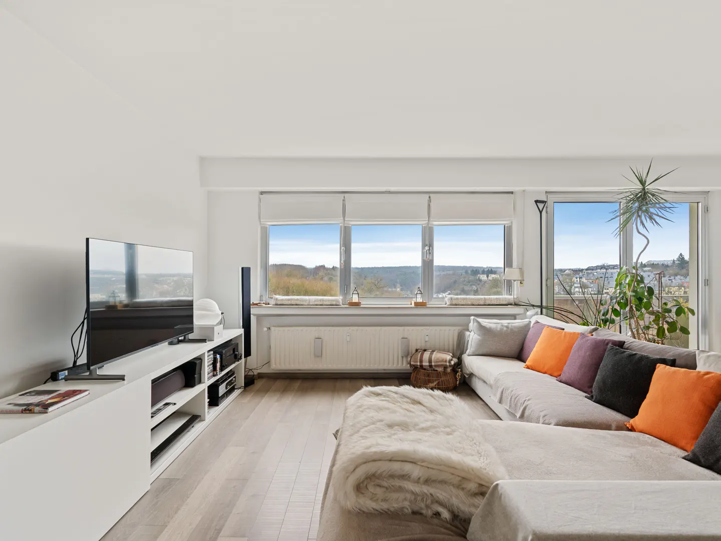 Bright living room with a large window view. A sectional sofa with colorful pillows faces a TV on a white console.