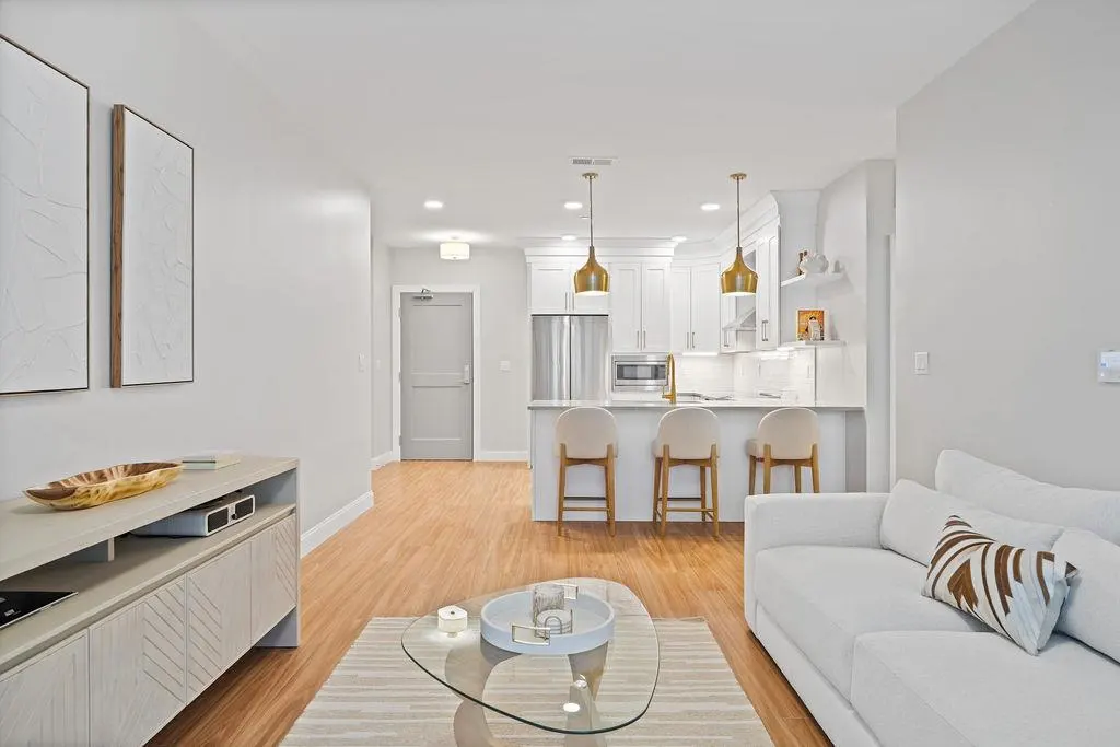 Bright, open-concept apartment with white sofa, glass table, and kitchen island with bar stools. Wood floors and modern decor.