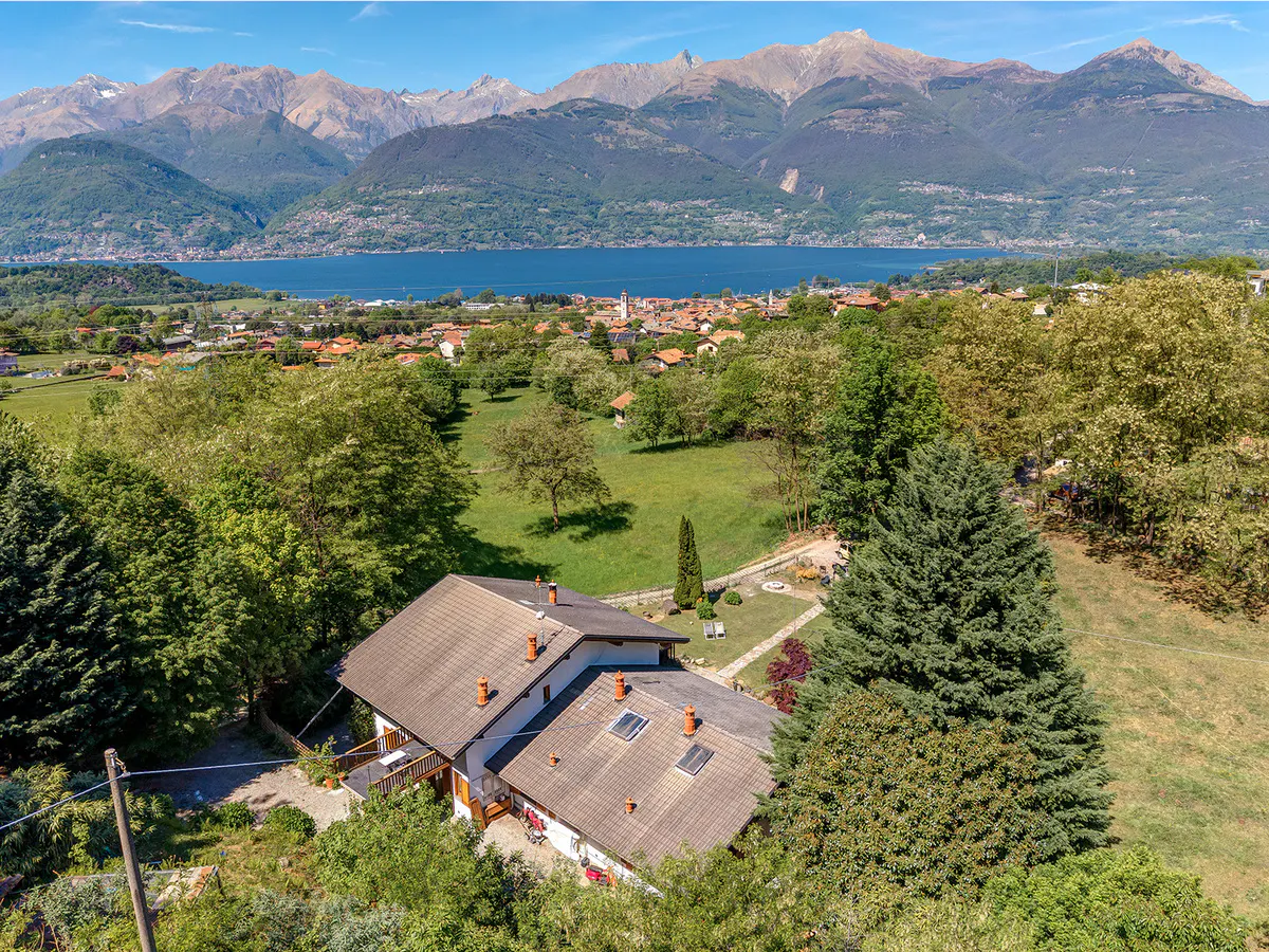 Aerial view of a house with a brown roof, surrounded by green trees, with a lake and mountains in the background.