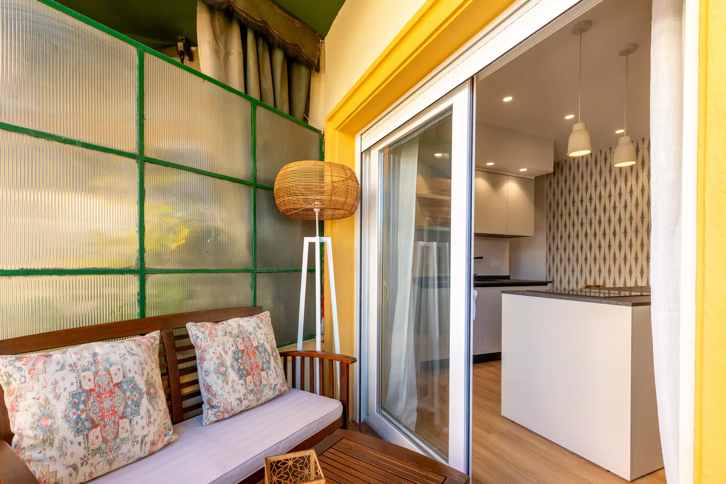 A bright balcony with a wooden bench, patterned pillows, and a woven lamp. Sliding glass doors open to a modern kitchen with pendant lights.