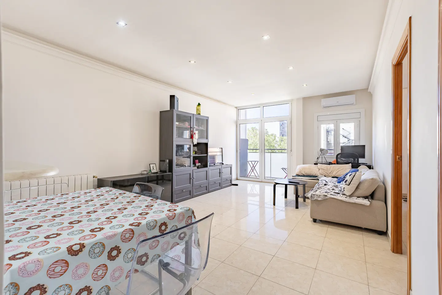 Bright living room with tile floors, white walls, and recessed lighting. A donut-patterned tablecloth covers a table with clear chairs. A gray entertainment center and beige sofa are also visible.