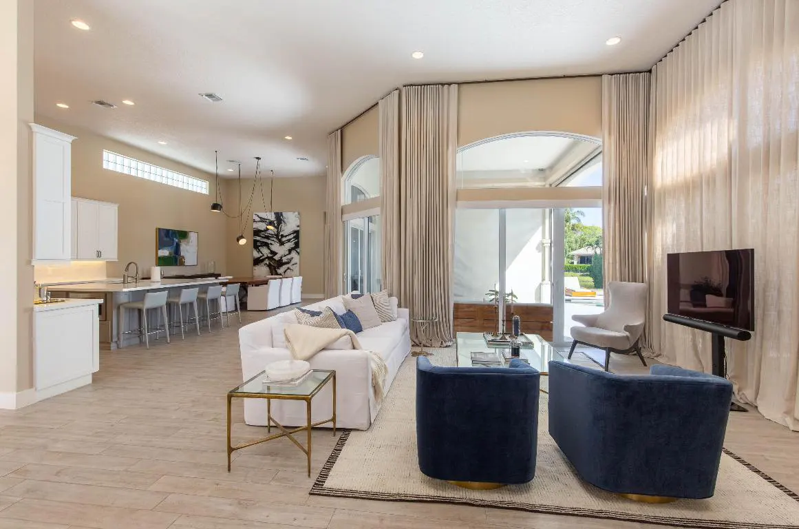 Open-concept living room with white sofa, blue chairs, and floor-to-ceiling curtains. Kitchen and dining area visible in the background.