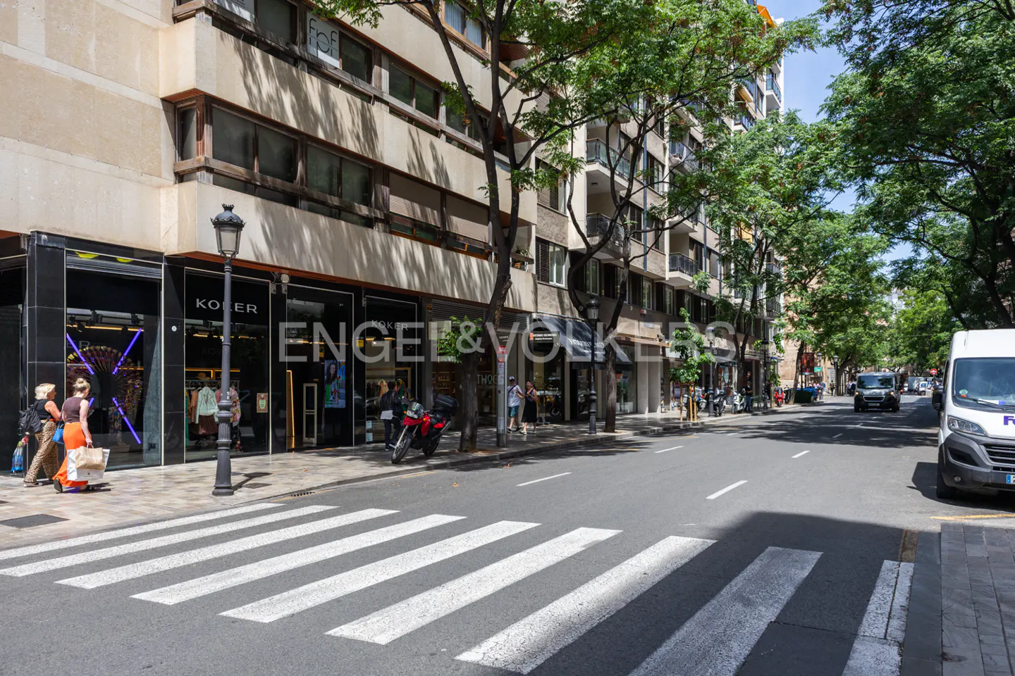 Street view of Engel & Volkers real estate office with shops, trees, crosswalk, and vehicles.