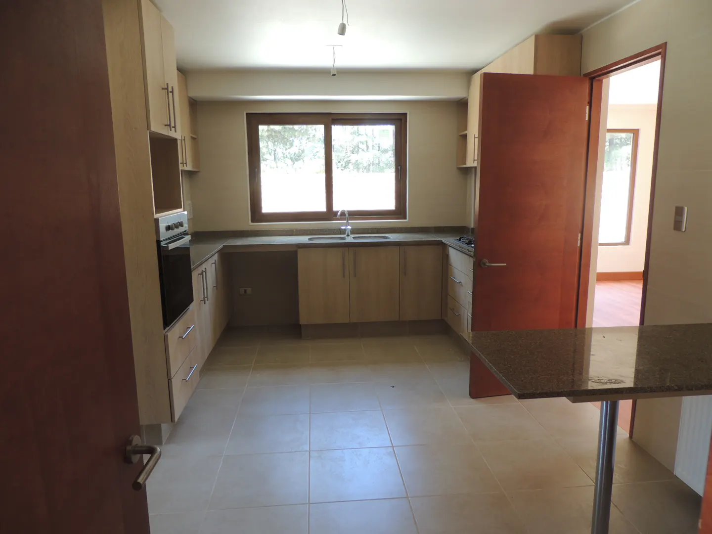 A bright kitchen with light wood cabinets, a dark countertop, and a window over the sink. A brown door is open to another room.