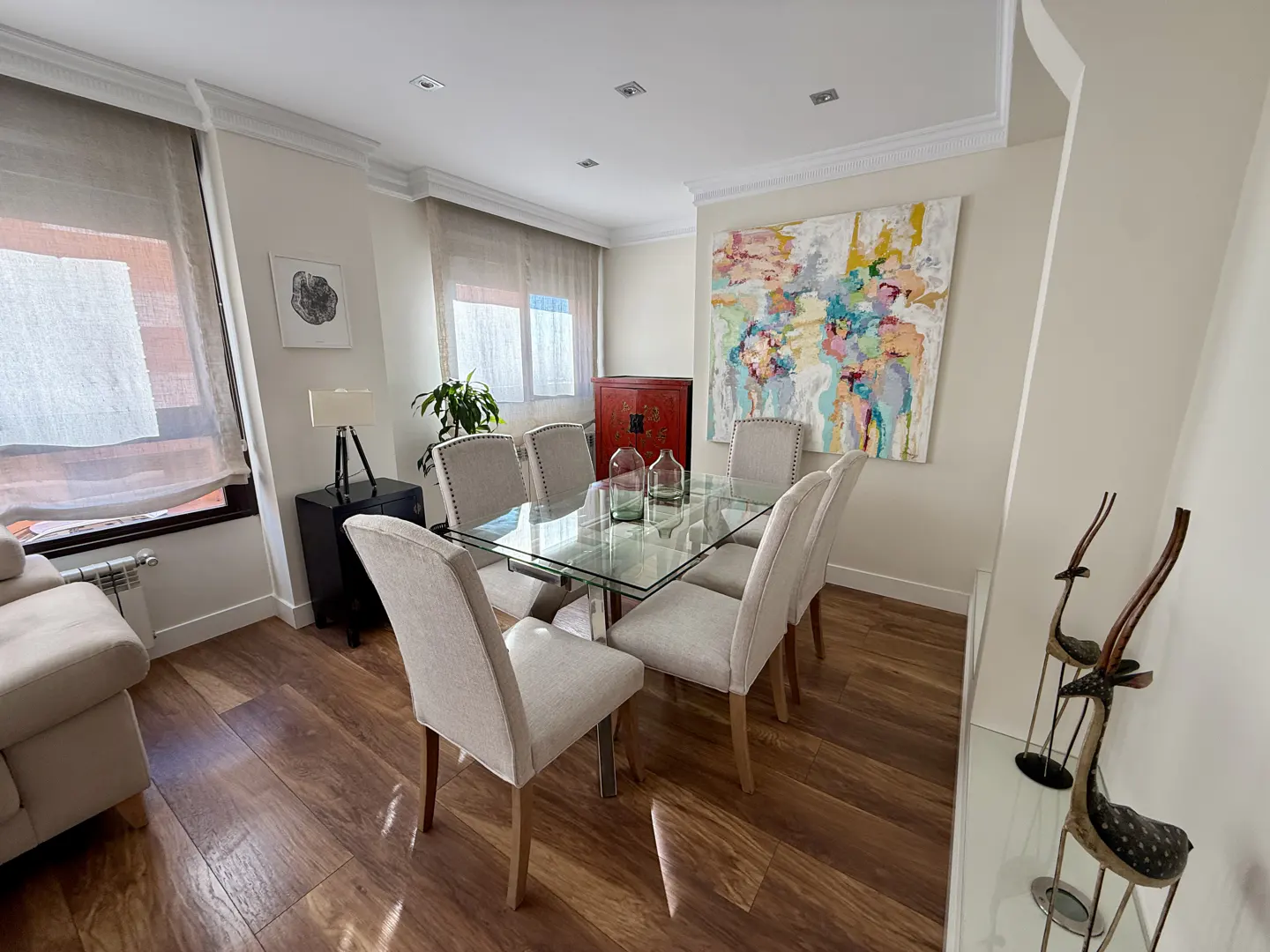 A dining room with a glass table, six chairs, and a colorful abstract painting on the wall.