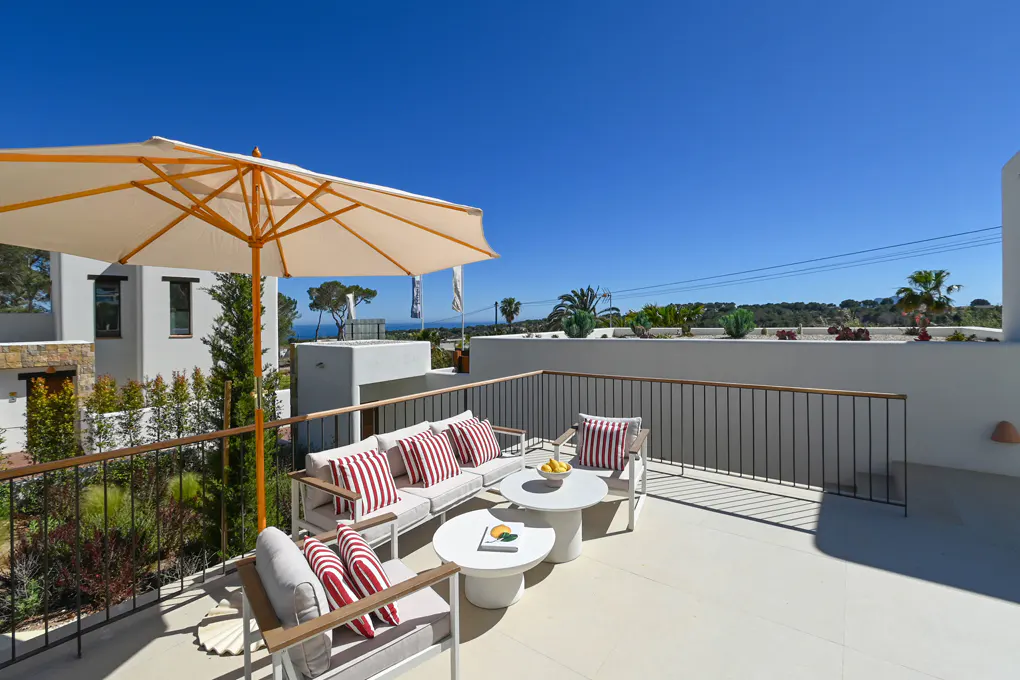 Outdoor patio with white furniture, red striped pillows, and a large umbrella. Railing surrounds the patio with a view of trees and a blue sky.