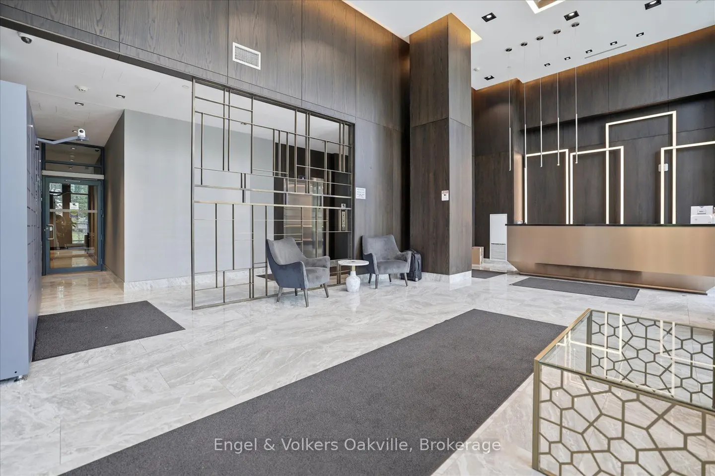 Modern lobby with marble floors, dark wood walls, a gold reception desk, two grey armchairs, and a geometric glass table.