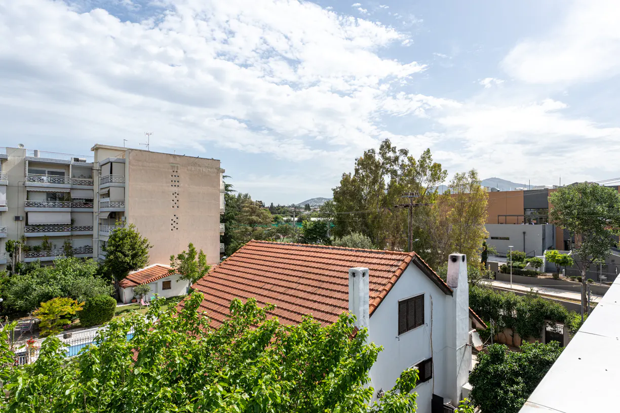 View of a white house with a red tile roof, surrounded by green trees, with buildings and a cloudy sky in the background.
