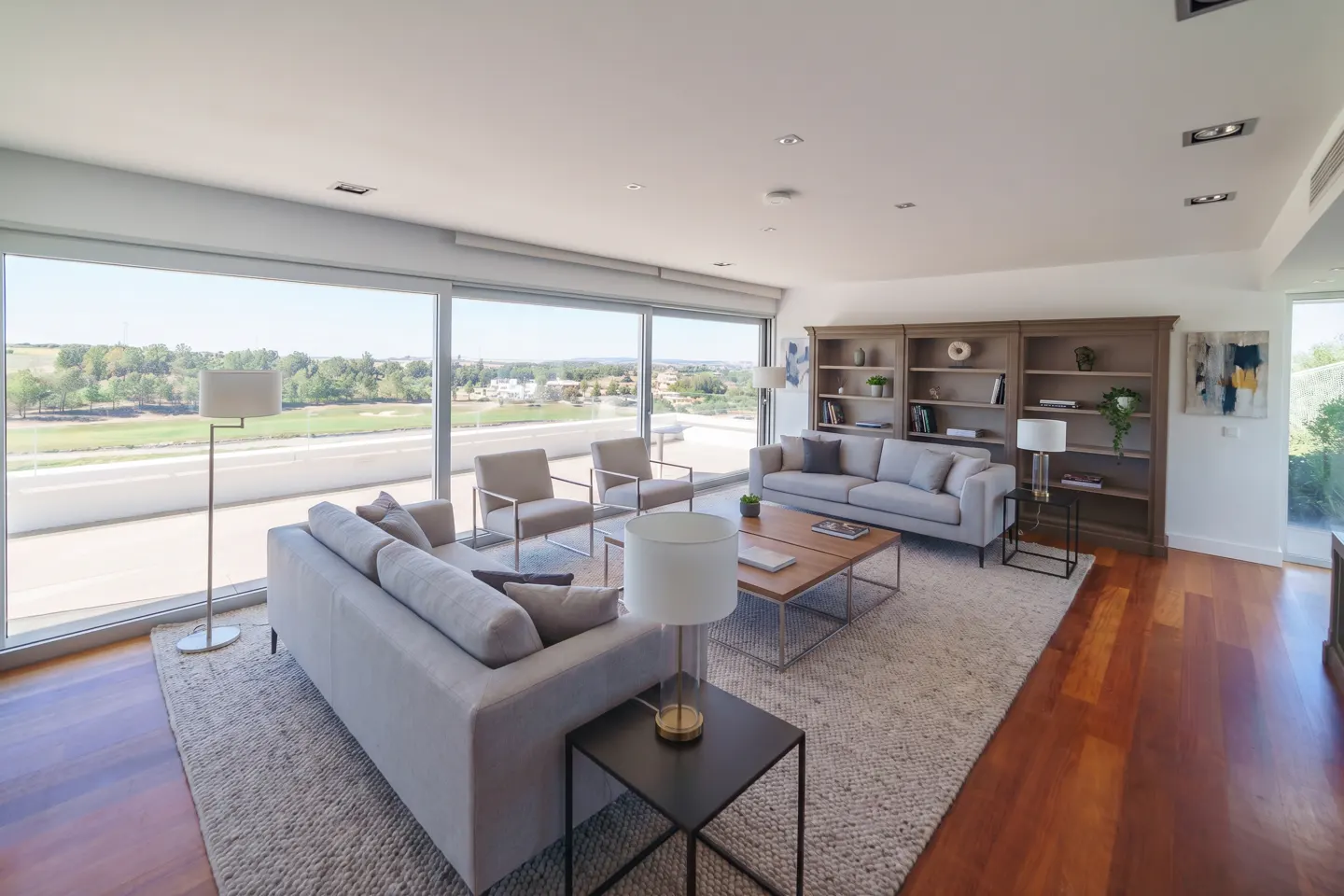 Bright living room with gray sofas, chairs, and a wood floor. Large windows offer a view of green fields. A bookshelf stands against the wall.