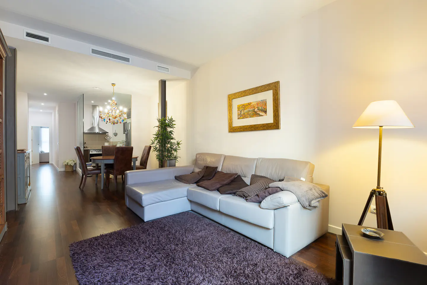 Living room with a gray sectional sofa, brown pillows, and a purple rug. A dining table and chandelier are in the background.
