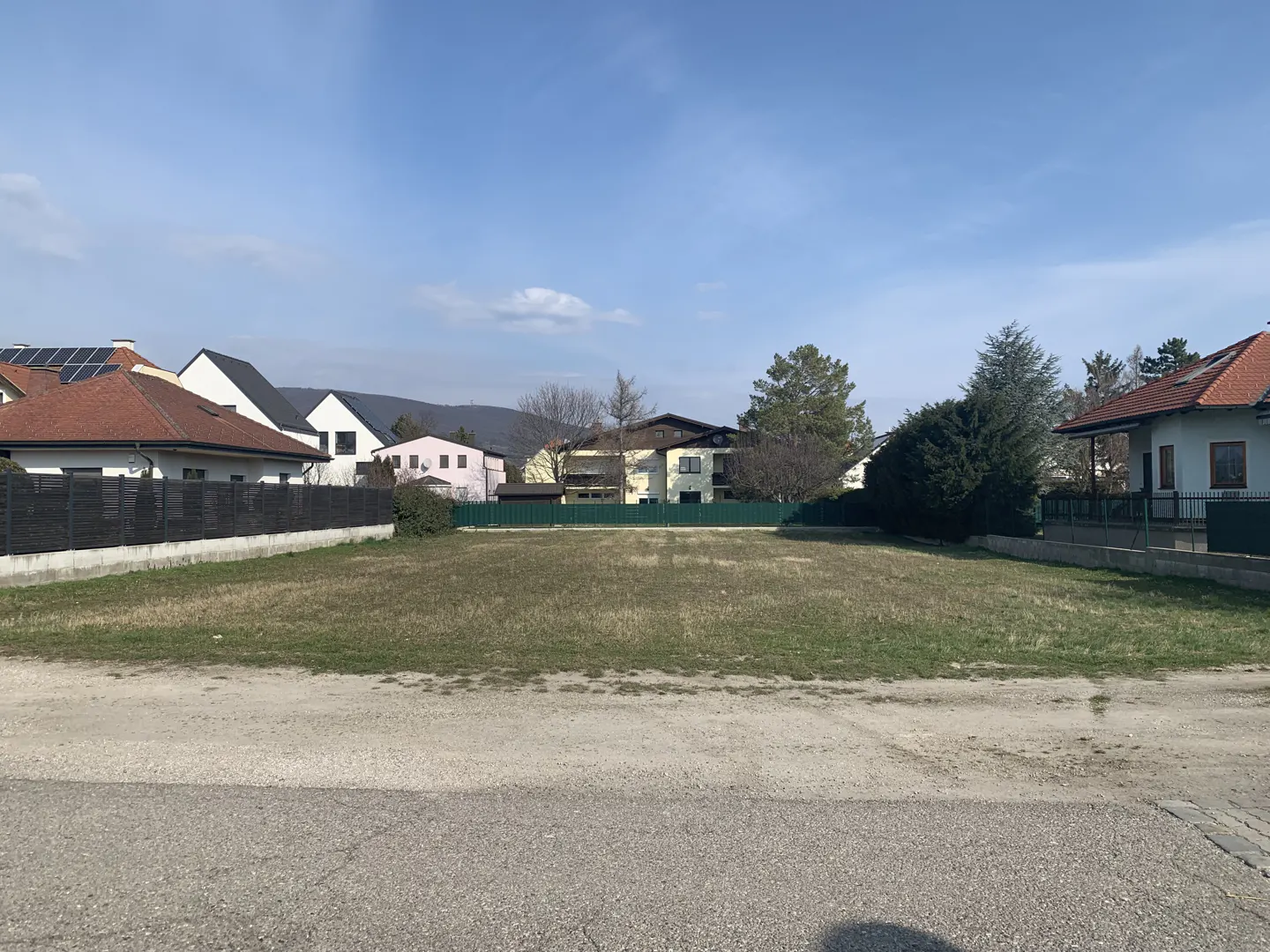 Vacant lot with dry grass, surrounded by houses and trees under a blue sky.