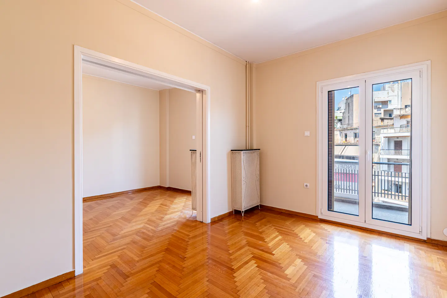 Empty room with beige walls, polished herringbone wood floors, a doorway to another room, and glass doors to a balcony.