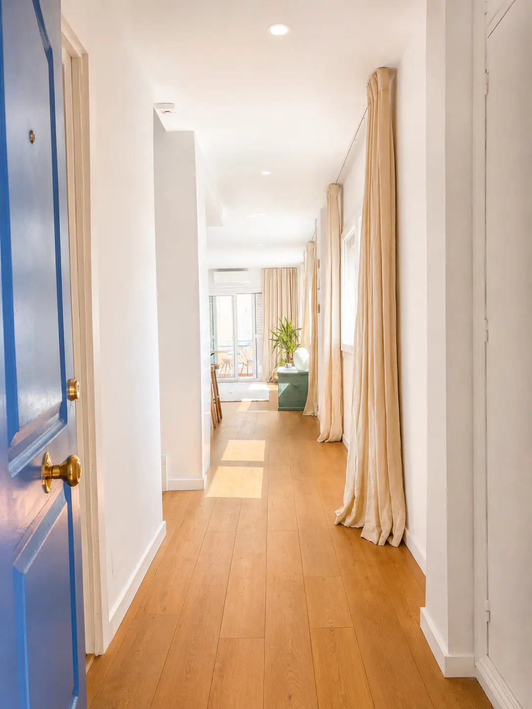 Bright hallway with wood floors, white walls, and a blue door ajar. Beige curtains hang near windows, leading to a sunlit room with a balcony.