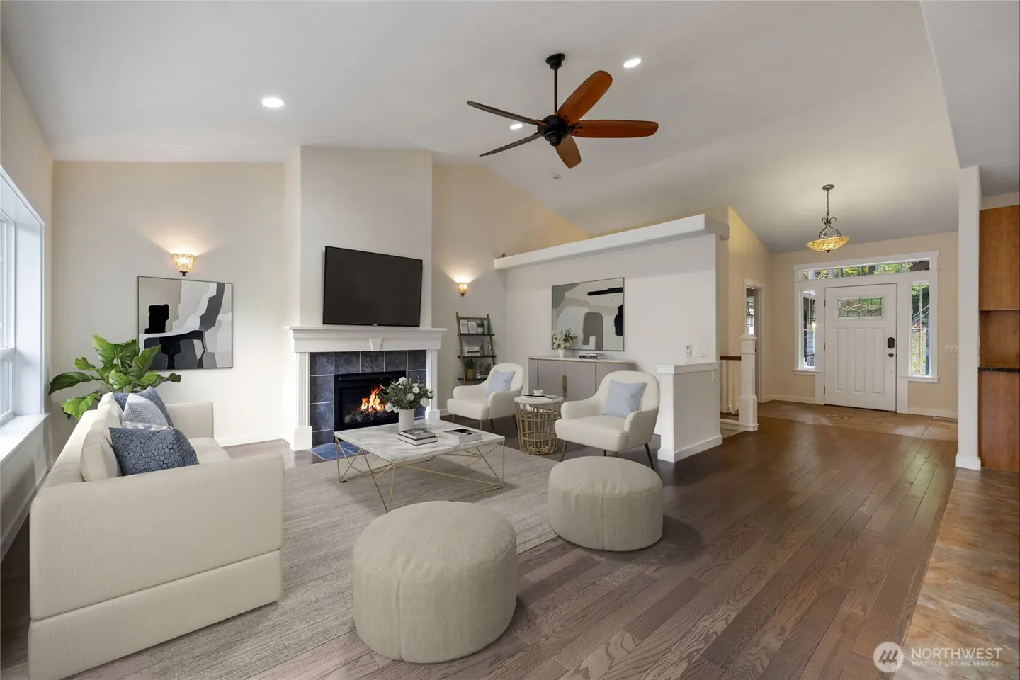 Bright living room with a white sofa, fireplace, TV, and hardwood floors. A ceiling fan and front door are also visible.