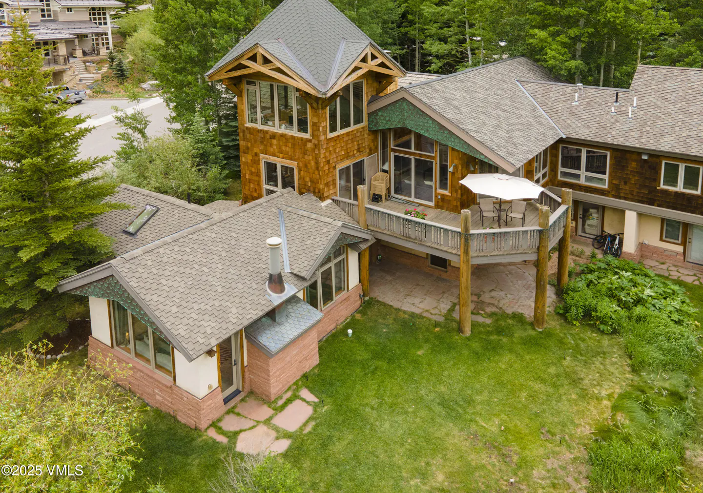 Aerial view of a multi-story brown wood and brick house with a deck, green lawn, and trees.