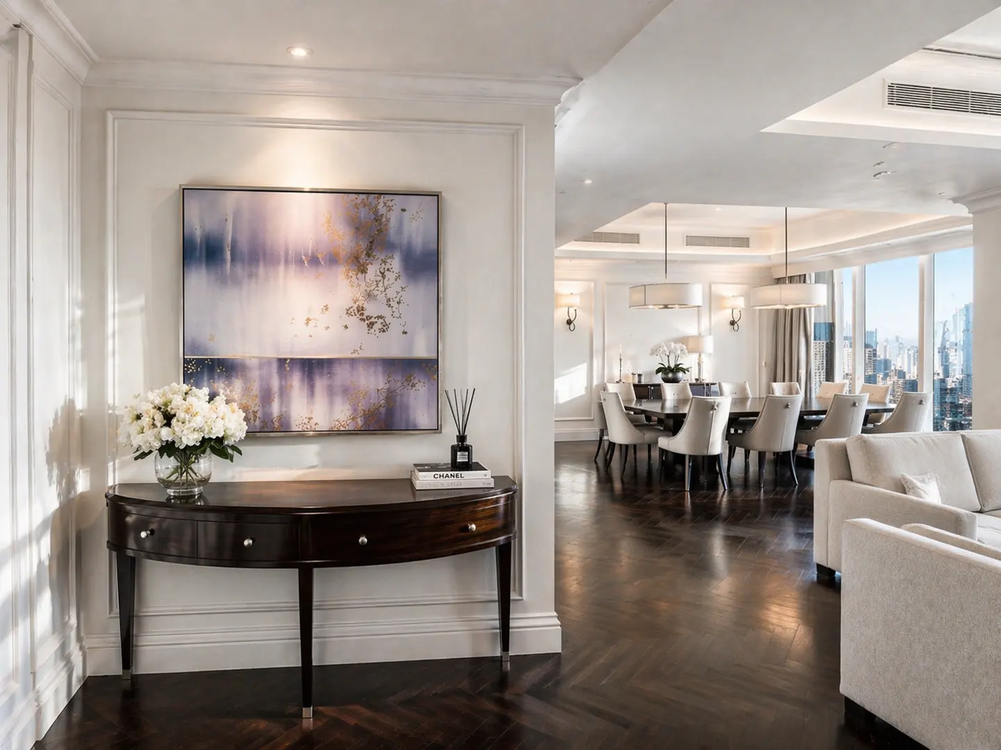 Elegant interior with dark wood floors, a half-moon table with flowers, and a painting. Dining area visible with city views.