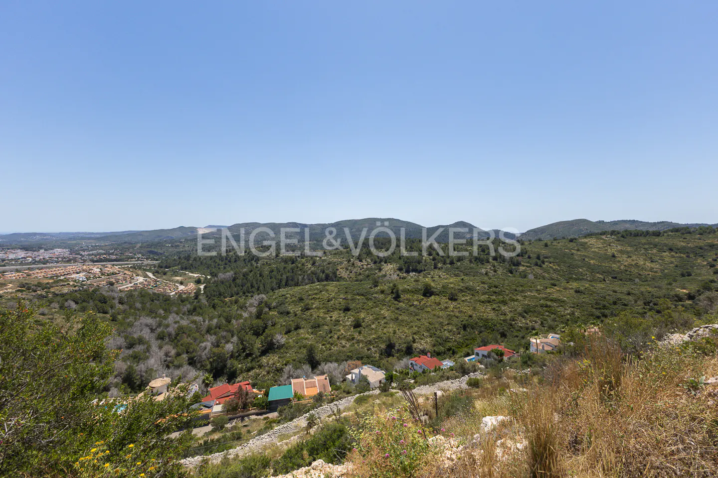 Hillsides with scattered homes and lush green trees under a clear blue sky. Distant mountains are visible.