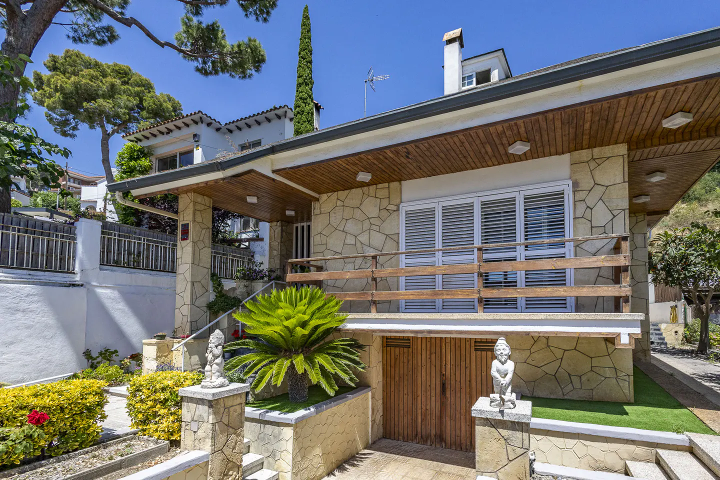 A stone-clad house with a wooden balcony, a palm plant, and two garden statues under a clear blue sky.