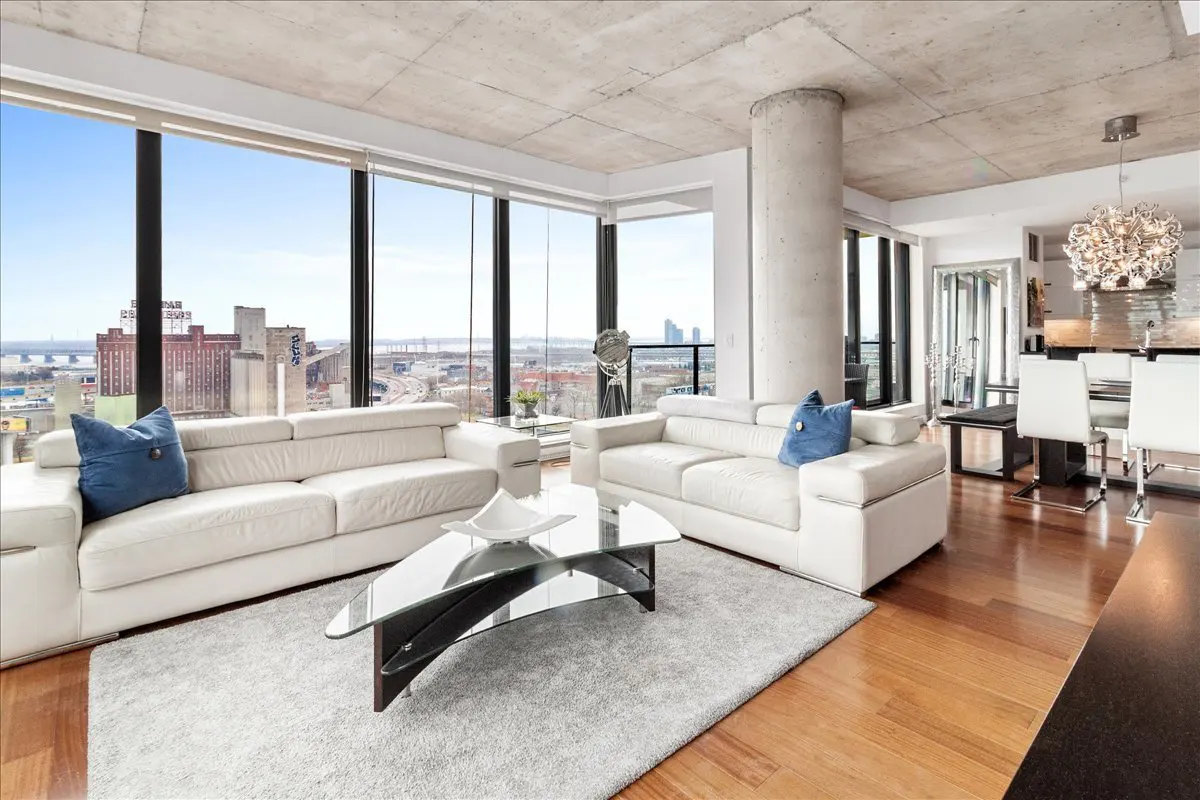 Bright living room with white sofas, blue pillows, and a glass coffee table on a gray rug. Large windows offer a city view.