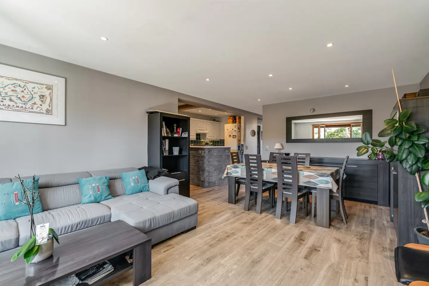 Open-concept living and dining area with gray sofa, blue pillows, and dark wood dining set. Kitchen visible in background.