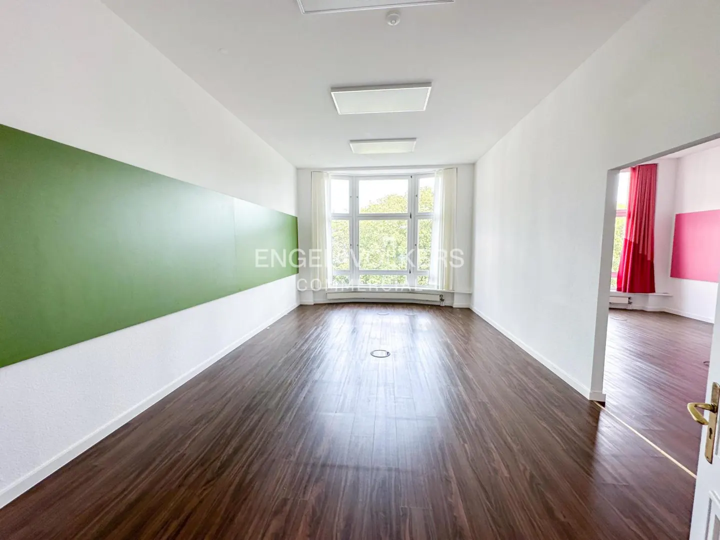 Bright, empty room with dark wood floors, white walls, and a green accent wall. A large window lets in natural light.