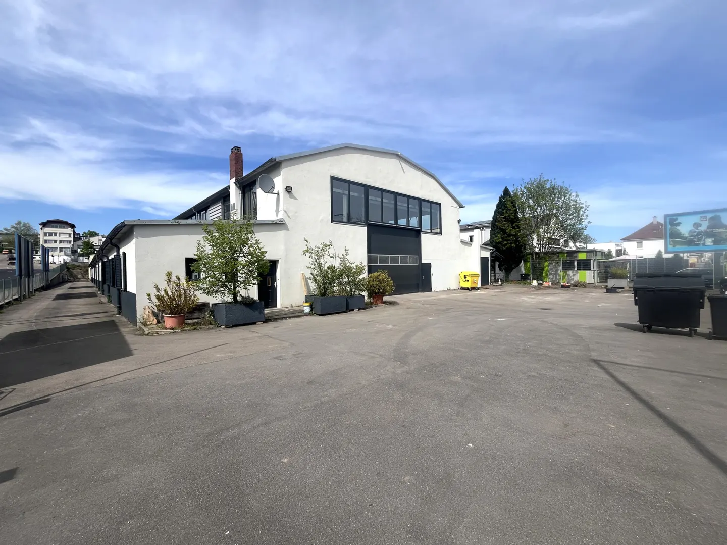 Exterior of a white commercial building with a large garage door and a paved parking lot. Trees and planters are in front of the building.