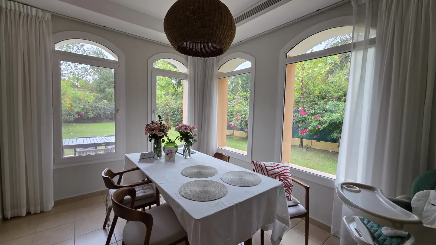 Dining area with a white table, floral centerpieces, and wooden chairs, surrounded by large windows overlooking a garden.