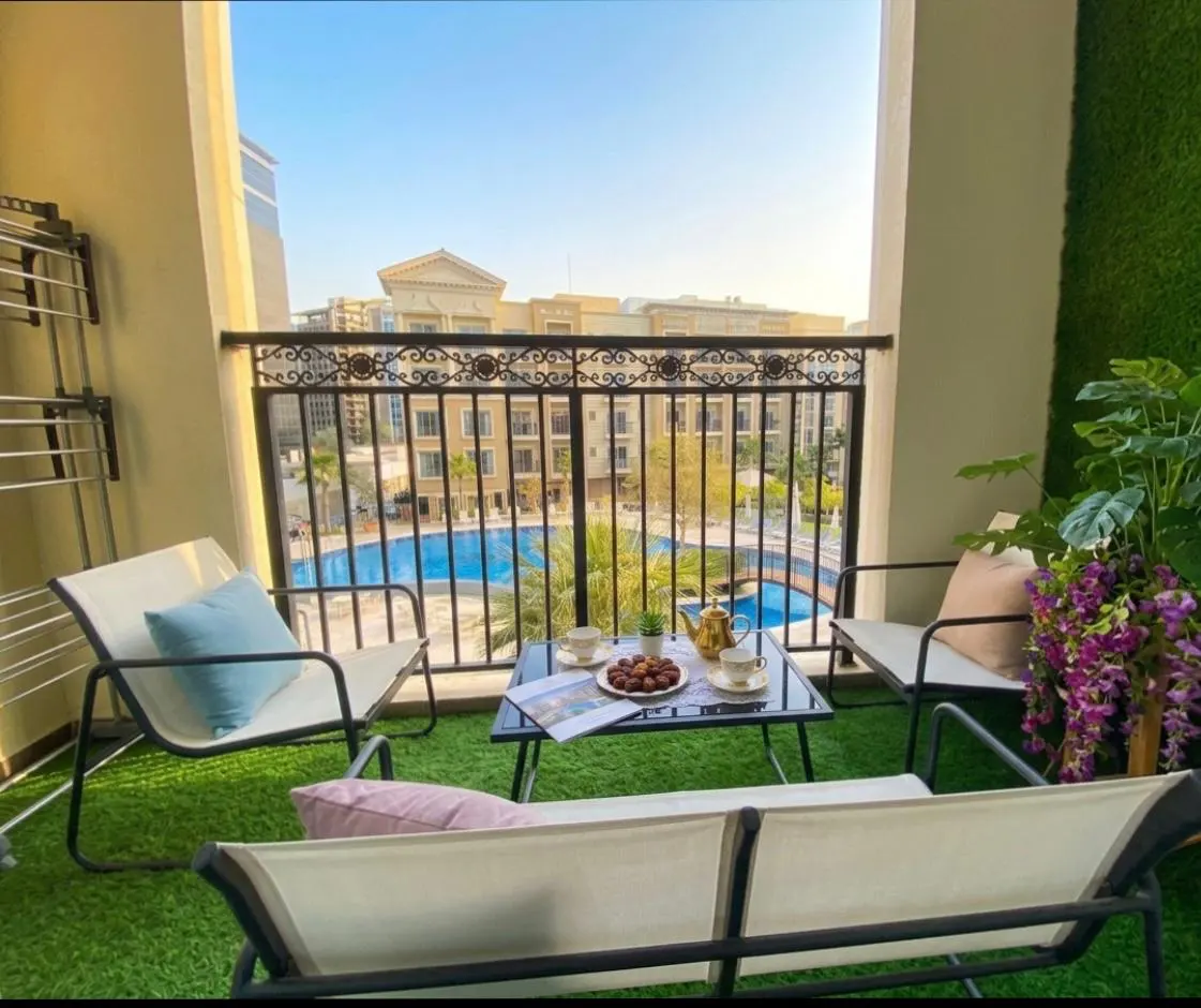 Balcony with artificial grass, seating, and a view of a pool. A table holds tea and dates. Plants add a touch of nature.
