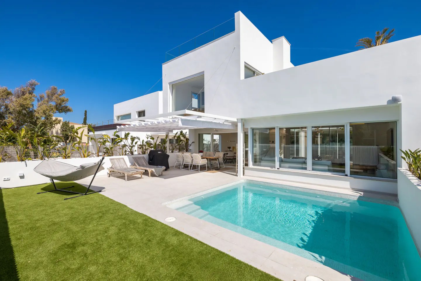 Modern white house with a pool, lawn, and patio furniture under a clear blue sky.
