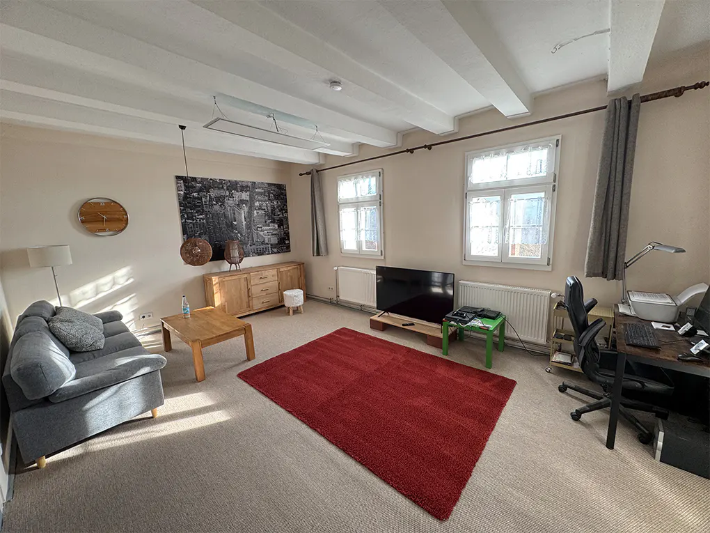 Living room with beige walls, white ceiling beams, and a red rug. A gray sofa, wooden furniture, and a black and white city print decorate the space.