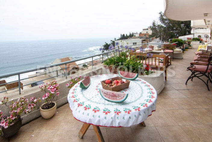 Balcony view with ocean backdrop. Table with floral cloth, fruit basket, and watermelon slices. Flowers and seating area visible.