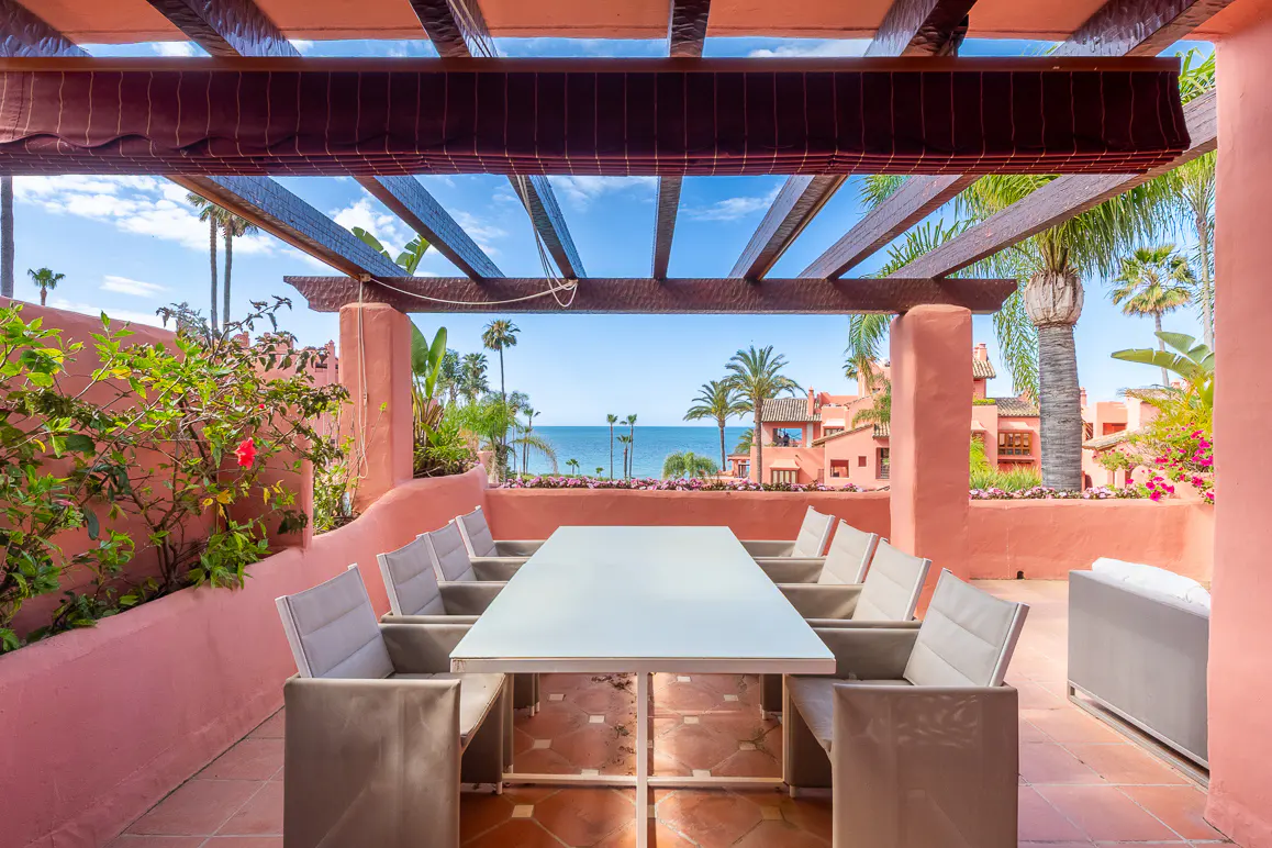 Outdoor dining area with a white table and gray chairs, under a pergola with a view of the ocean and palm trees.