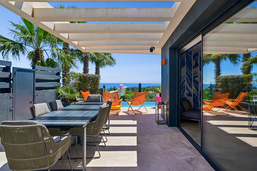 Modern patio with a dining table, wicker chairs, and orange lounge chairs overlooking a swimming pool and the ocean.