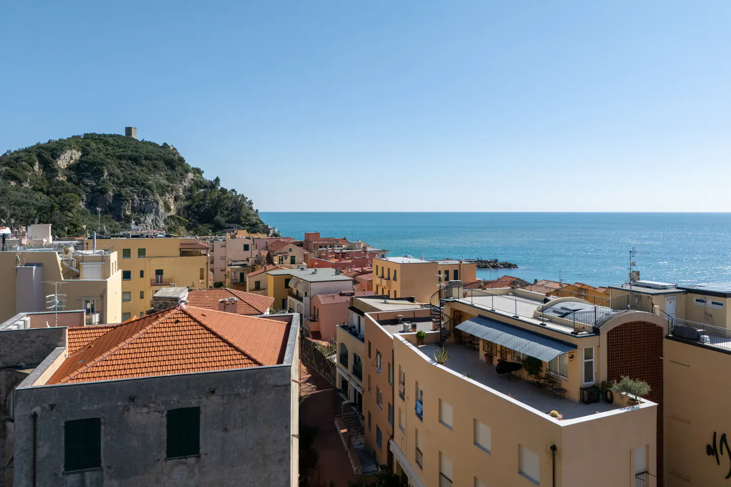 Coastal view of Lerici, Italy, featuring colorful buildings with red tile roofs, a green hillside with a tower, and the blue Ligurian Sea.