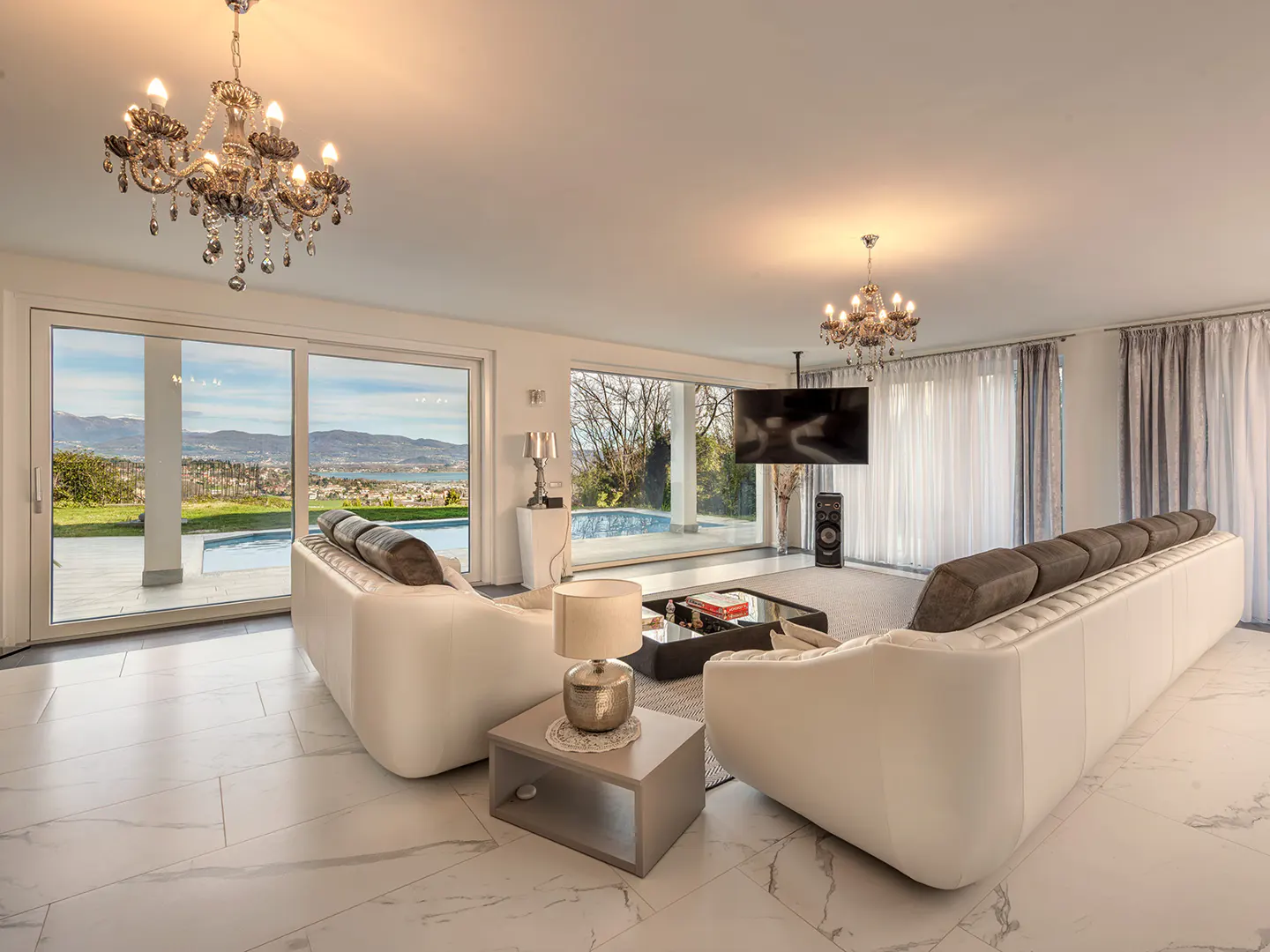 Bright living room with white marble floors, white sofas, and chandeliers. Large windows overlook a pool and landscape.