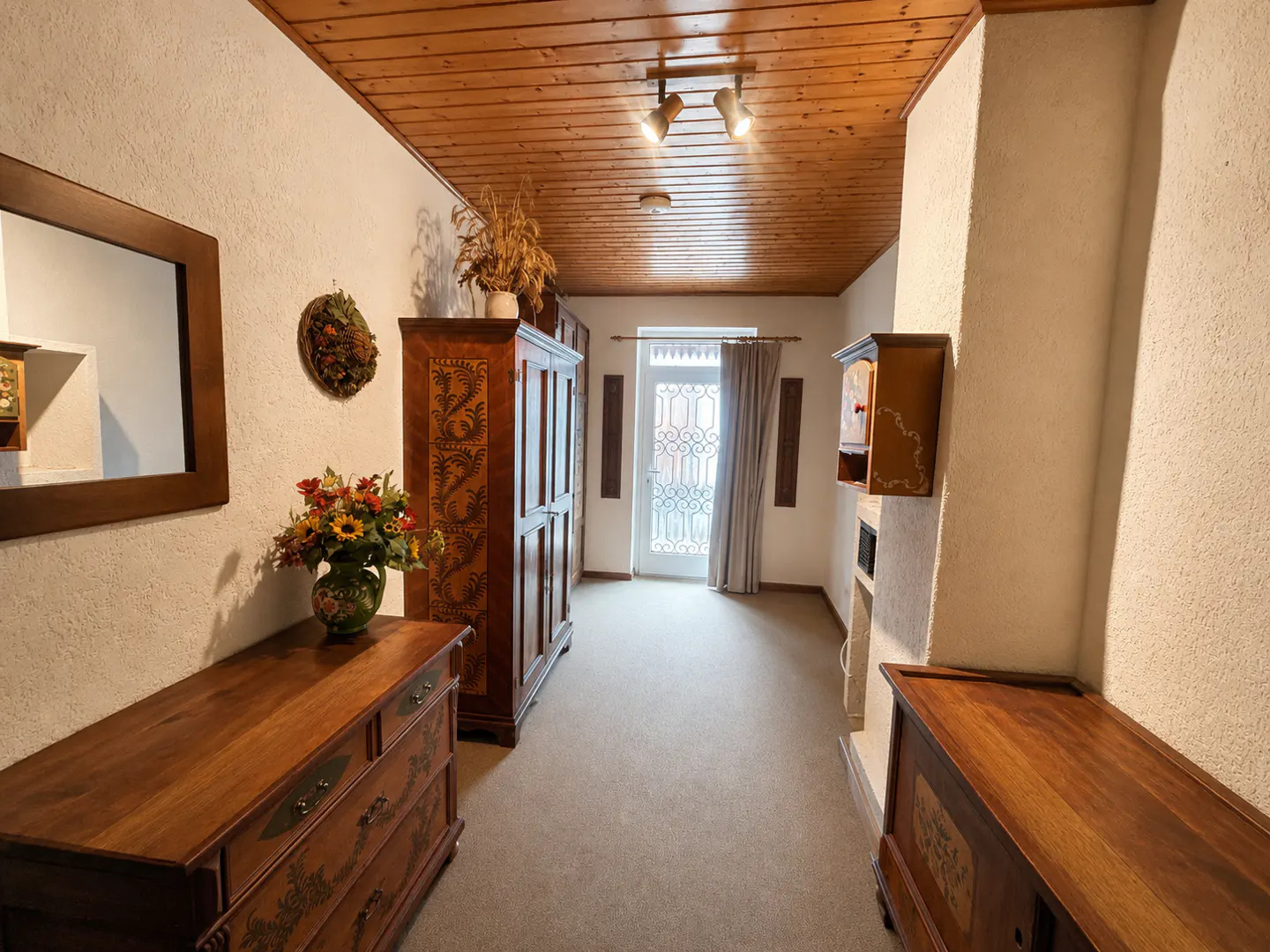 Hallway with wooden ceiling, white walls, and beige carpet. A wooden dresser with flowers, a mirror, and a wardrobe are on the left. A door is at the end.