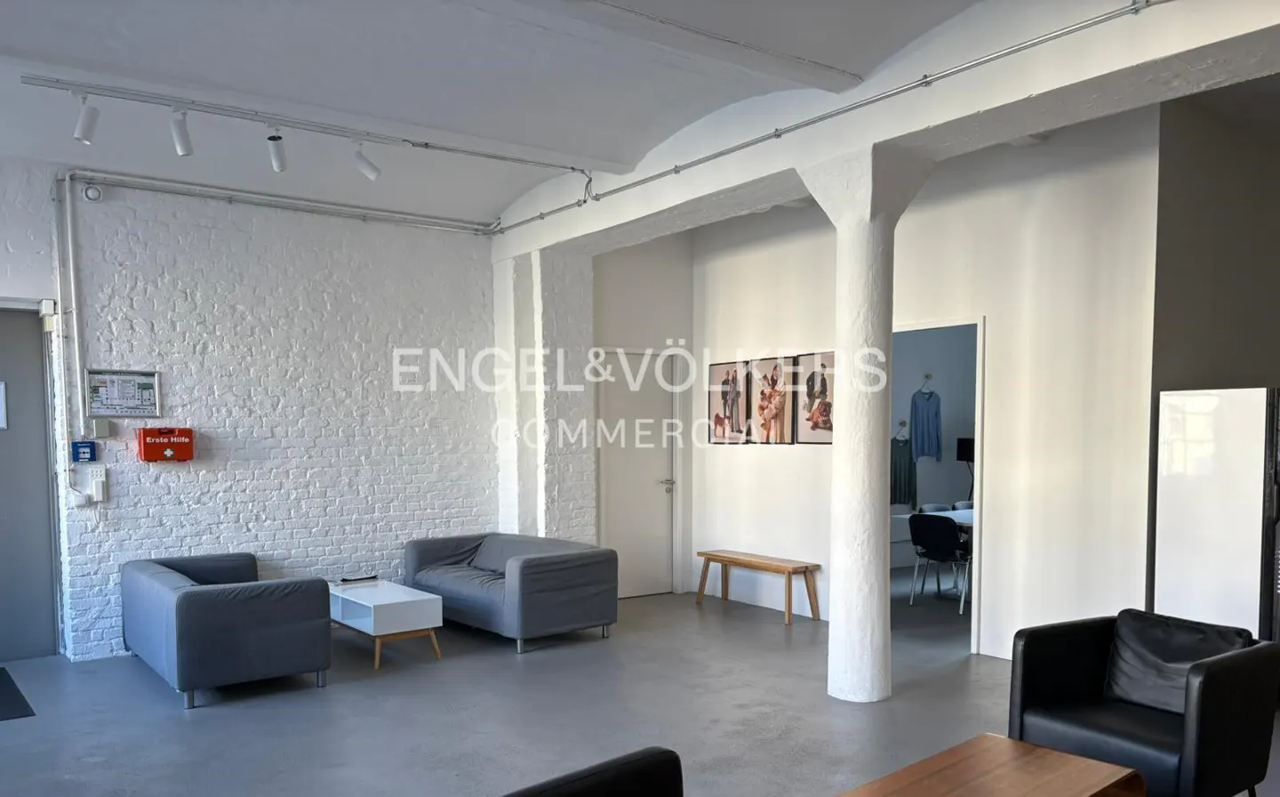 Bright, modern office space with gray sofas, white brick walls, and concrete floors. A doorway leads to another office area.