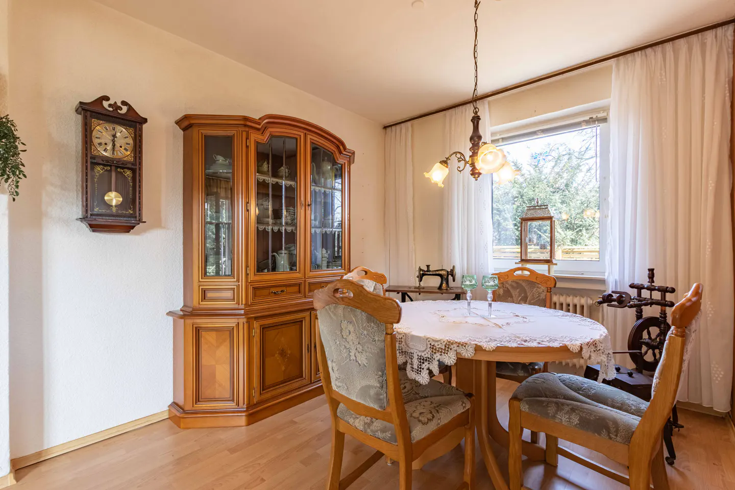 Dining room with a wooden hutch, round table with lace cloth, vintage wall clock, and antique sewing machine by a window.