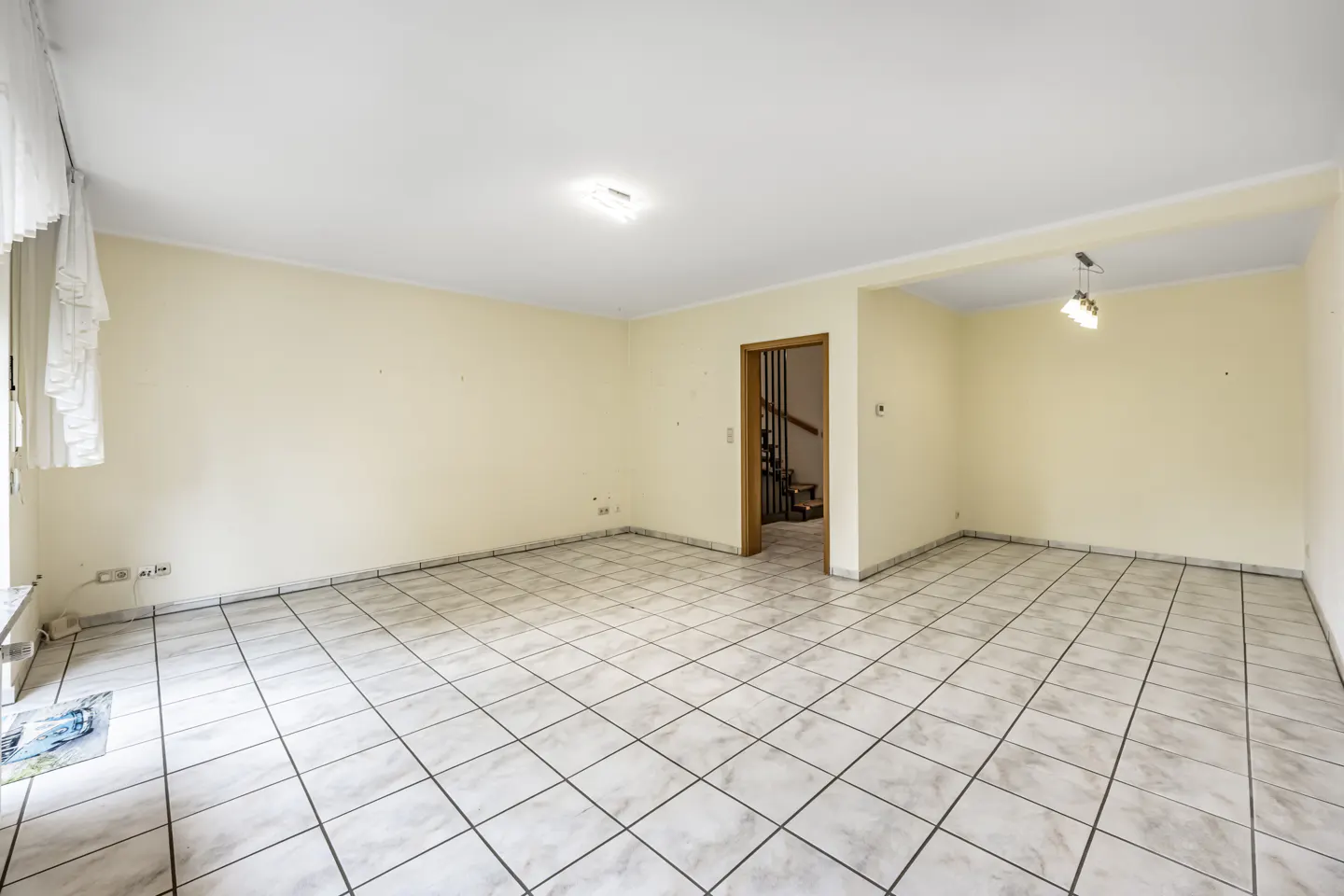 Empty, spacious room with cream walls, white tiled floors, and a doorway leading to a staircase.