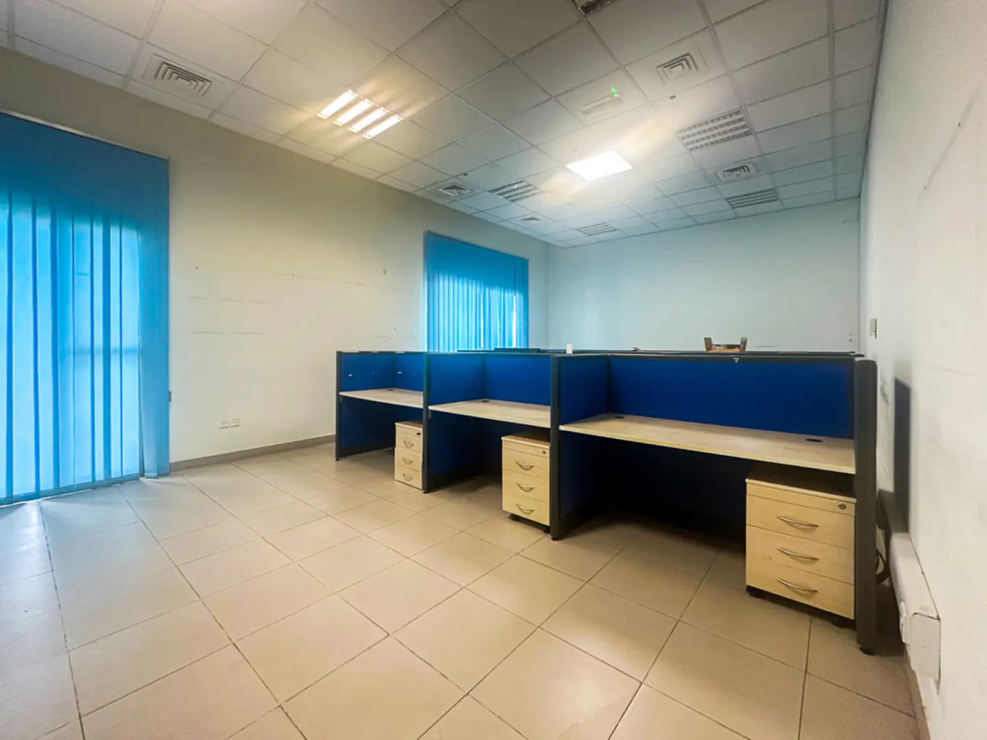 Empty office space with blue cubicles, beige desks, and file cabinets. Blue vertical blinds cover the windows. Tiled floor and fluorescent lighting.