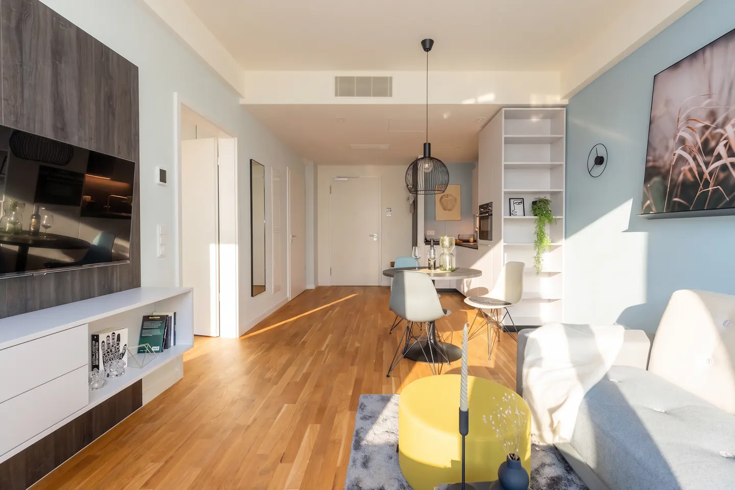 Bright, modern apartment interior with wood floors, a gray sofa, and a yellow ottoman. A round table with chairs sits under a black pendant light.