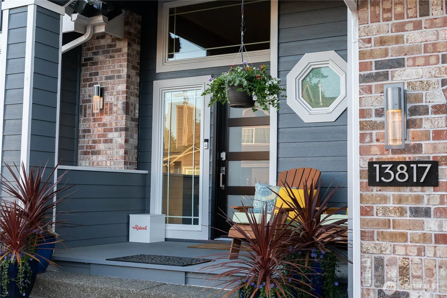 Front porch of a home with dark blue siding, brick accents, a modern glass door, hanging flowers, and house number 13817.